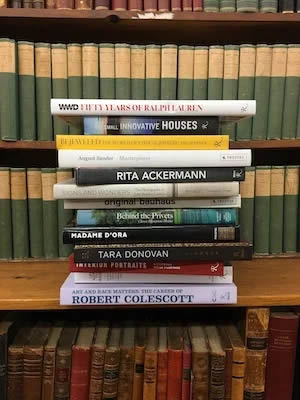 A stack of books on a wooden shelf, with titles related to art, architecture, and design, against a backdrop of green and brown spines.